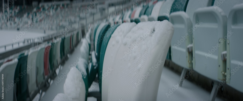 Fototapeta premium Empty outdoor football soccer stadium seats covered with snow in winter, light snowfall. Shot with 2x anamorphic lens