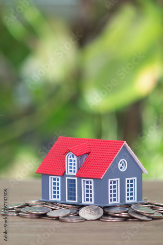 A small house model is placed on a pile of dollar coins on the table in front of the blurred greenery background