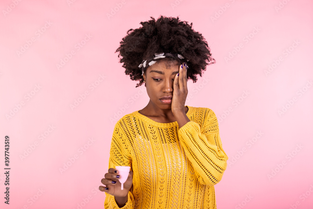 Pensive African American woman holds effective safe affordable menstrual product, good protection from leakage, chooses reusable menstrual cup, thinks how to use it, blank empty space on pink wall Pensive African American woman holds effective safe affordable menstrual product, good protection from leakage, chooses reusable menstrual cup, thinks how to use it, blank empty space on pink wall