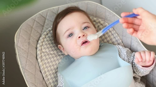 Cute hungry toddler boy impatiently waiting for feeding and mom feeds him porridge from a spoon side view photo