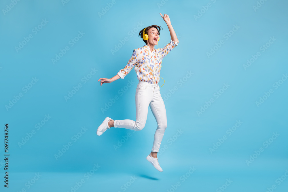 Full size photo of young girl happy excited jump listen music earphones show finger empty space isolated over blue color background Full size photo of young girl happy excited jump listen music earphones show finger empty space isolated over blue color background