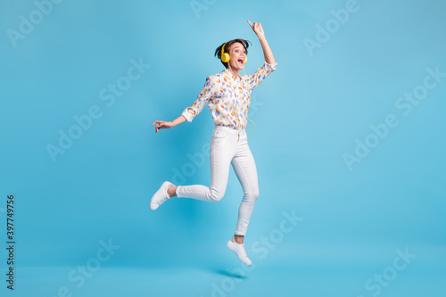 Full size photo of young girl happy excited jump listen music earphones show finger empty space isolated over blue color background