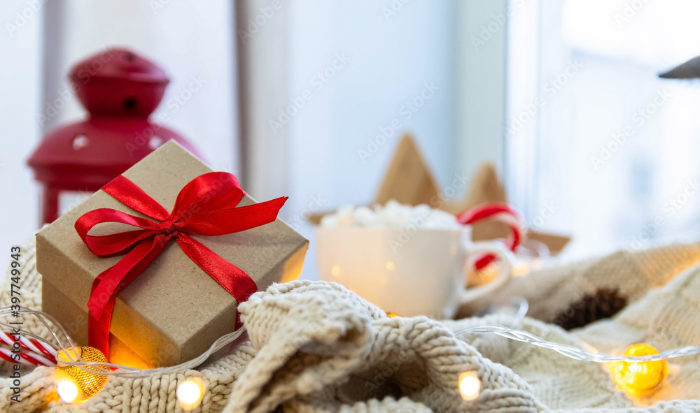Gift with a red ribbon on a white knitted blanket. New Year, garlands, gifts, a cup and marshmallows - an atmosphere of warmth, comfort and magic. Gift with a red ribbon on a white knitted blanket. New Year, garlands, gifts, a cup and marshmallows - an atmosphere of warmth, comfort and magic.
