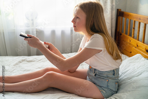 a 13 year old girl is lying on the bed and holding the TV remote in the bedroom near the window
