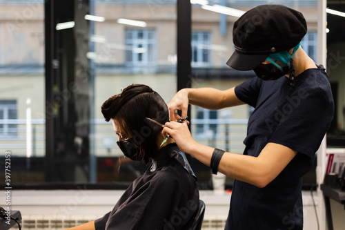 A professional girl hairdresser makes a client haircut. The girl is sitting in a mask in beauty the salon. Social distance. Protective measures. Both woman wearing face masks.
  