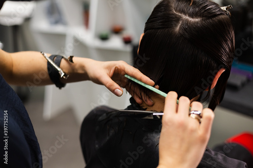 A professional girl hairdresser makes a client haircut. The girl is sitting in a mask in beauty the salon. Social distance. Protective measures. Both woman wearing face masks. 