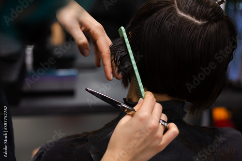 A professional girl hairdresser in face mask makes a client trendy stylish short hair cut. The girl is sitting in stylish beauty salon. Social distance. Protective measures.   