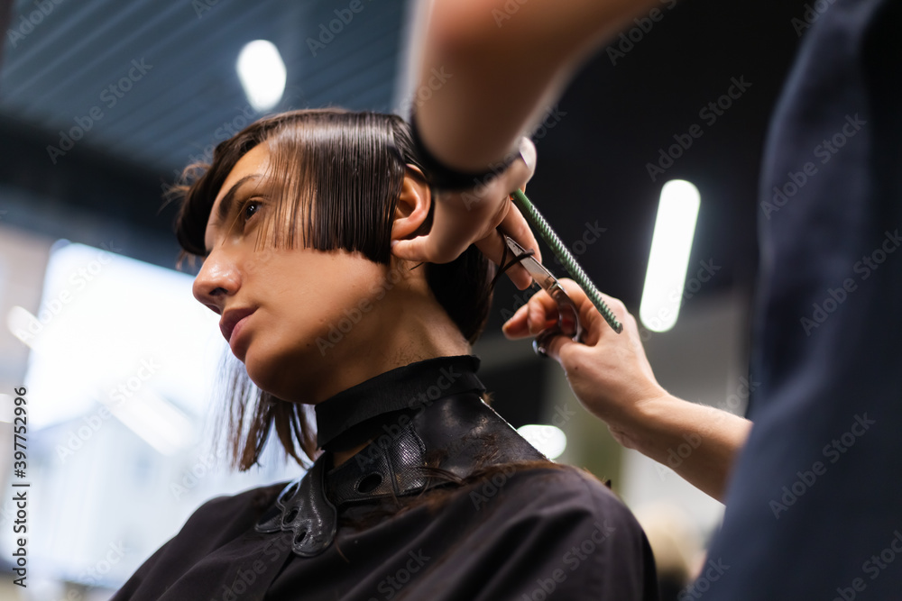 A professional girl hairdresser in face mask makes a client trendy stylish short hair cut. The girl is sitting in stylish beauty salon. Social distance. Protective measures.
  