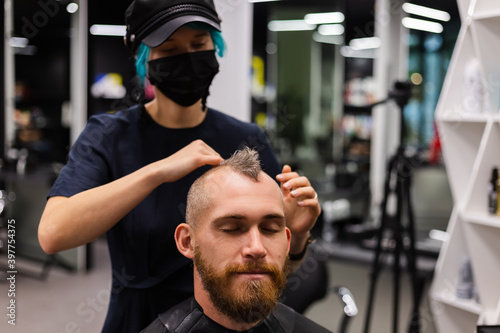 Professional hairdresser wearing protective face mask, make haircut for european bearded brutal man in beauty salon. 