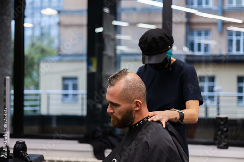 Professional hairdresser wearing protective face mask, make haircut for european bearded brutal man in beauty salon.
 