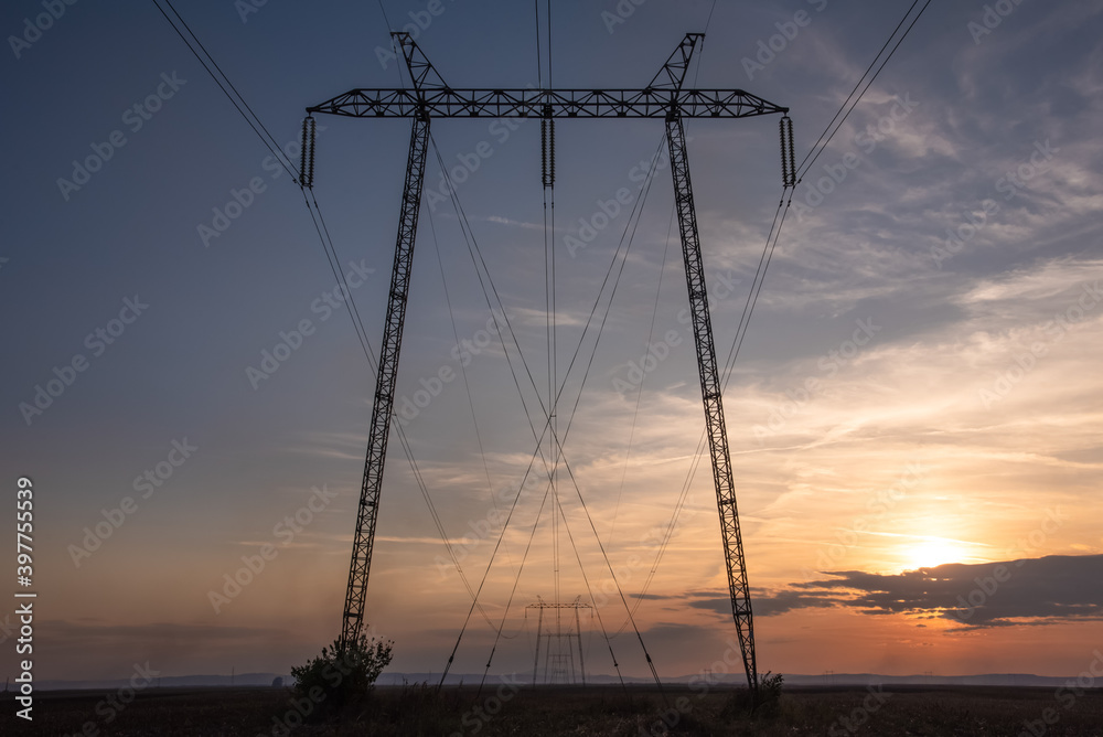 Silhouette of High voltage transmission lines and towers runing trough lawn and grass field in sunset with beautiful colorful sky in the background.