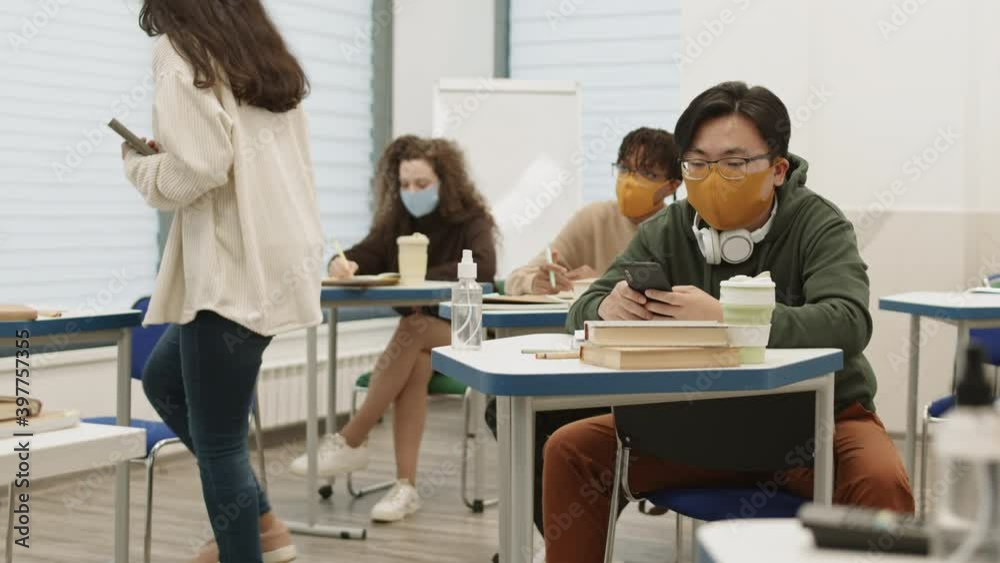 Diverse students sitting by desks in classroom on break, Asian guy in glasses using smartphone, Mixed-Race girl entering, taking her place. Students wearing masks, learning during pandemic Diverse students sitting by desks in classroom on break, Asian guy in glasses using smartphone, Mixed-Race girl entering, taking her place. Students wearing masks, learning during pandemic