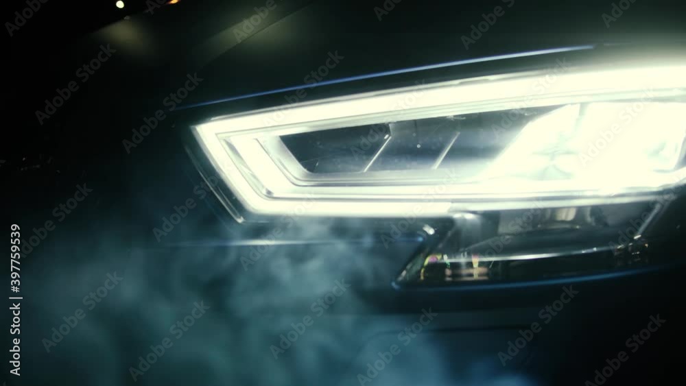 Close up view on the switched on headlights of the black car in the dark. Smoke slowly hovers around. Beautiful light in garage. Close up view on the switched on headlights of the black car in the dark. Smoke slowly hovers around. Beautiful light in garage.