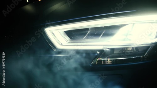 Close up view on the switched on headlights of the black car in the dark. Smoke slowly hovers around. Beautiful light in garage.