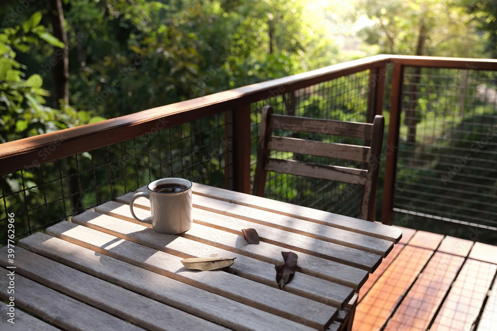 a coffee cup and many dried leaves on wooden table and chair at balcony with green trees in background in afternoon