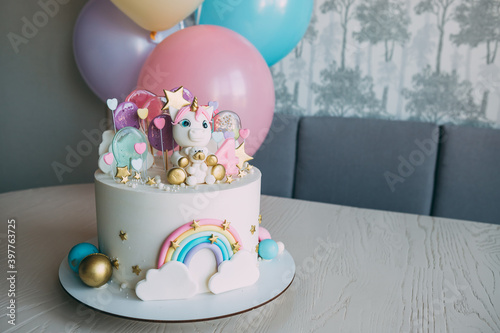 Birthday cake to a girl with a unicorn and balloons..Cake for a girl of gentle pastel tones, decorated with unicorn figures with a bear and rainbow, lollipops, on the background - colorful balls