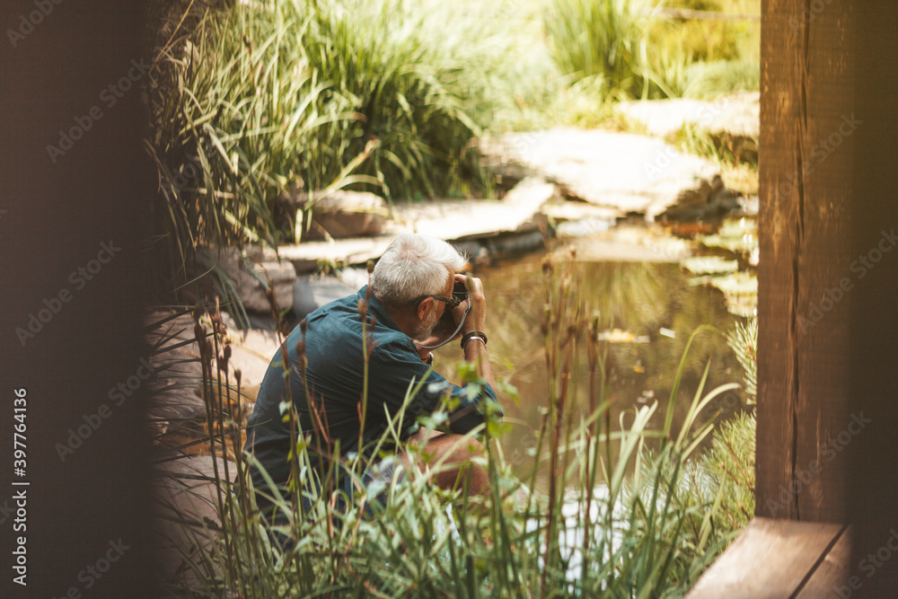 Fototapeta premium Elderly man taking pictures of water as a hobby. Leisure in old age. Relaxing at home in the garden alone.