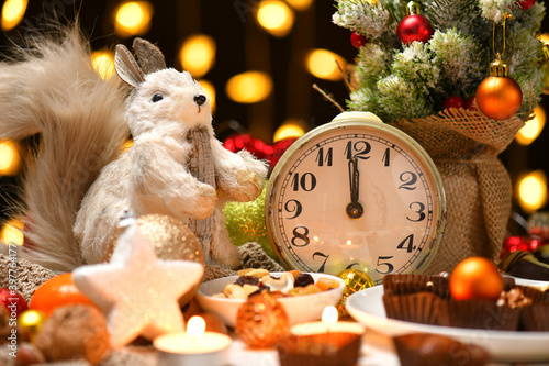 christmas or holiday decoration background - small fir tree, retro alarm clock shows midnight and sweet food. New year still life with xmas lights