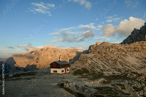 A capture of the Tre Cime di Lavaredo cottage  Drei Zinnenhuette  in Italian Dolomites during the golden hour. The cottage has red decorative elements. There are high Alpine peaks around. Shelter