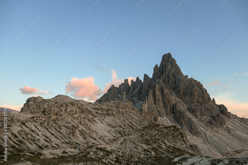 A capture of high and sharp peaks of Dolomites in Italy during the golden hour. The sky is full of soft clouds. Lots of lose stones and pebbles. Raw and desolated landscape. Serenity and calmness