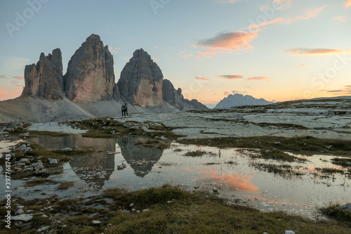 A couple enjoying the sunset over the Tre Cime di Lavaredo (Drei Zinnen) mountains in Italian Dolomites. The peaks reflect in a paddle. The mountains are surrounded with orange and pink clouds. Love