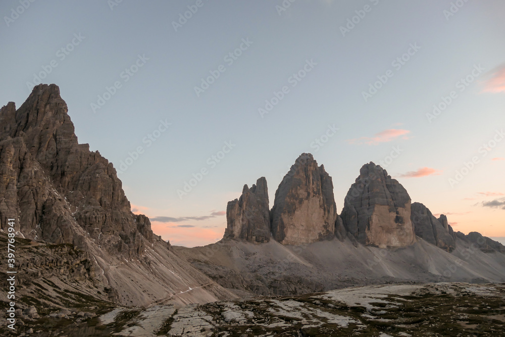 A panoramic capture of the sunset above Tre Cime di Lavaredo (Drei Zinnen) and surrounding mountains in Italian Dolomites. The mountains are surrounded by pink and orange clouds. Golden hour. Serenity