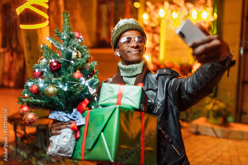 Portrait of an African American young man smiling taking a selfie with Christmas presents and a Christmas tree. In anticipation of the Christmas holidays. Christmas concept.