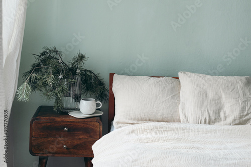 Christmas still life. Cup of coffee, retro wooden bedside table. Pine tree branches and silver stars ornaments in glass vase. Bed with beige cotton muslin beddings. Blank sage green wall background. photo