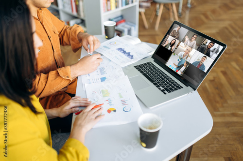 Online video chat. Laptop monitor view with multiracial business colleagues discussing about financial graphs, man and woman sitting on their workplace during video conference