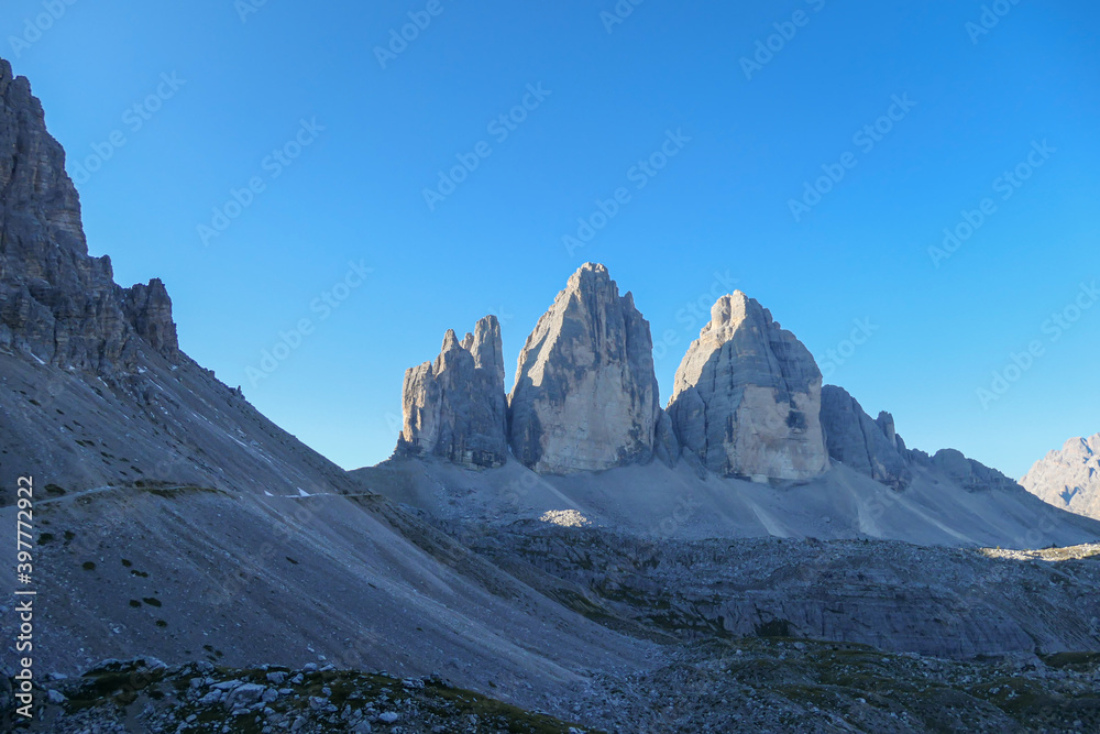 Panoramic view on the first sunbeams reaching the top of the Tre Cime di Lavaredo (Drei Zinnen), Italian Dolomites. There is a narrow path leading to the peak. New day beginning in the high mountains