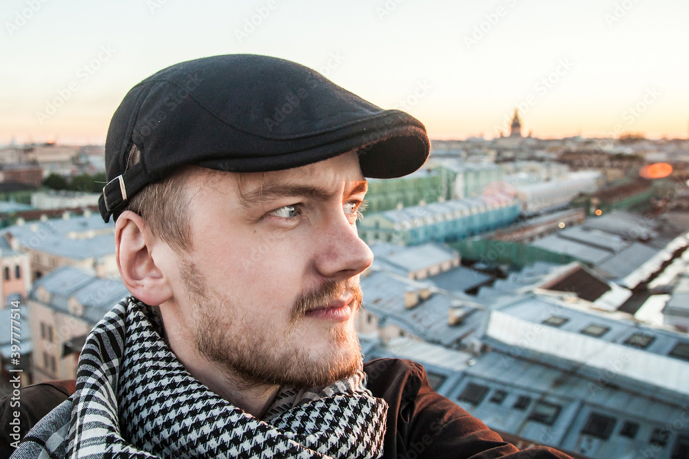 Portrait of smiling guy with beard and blue eyes in black cap on a sunset rooftop background
