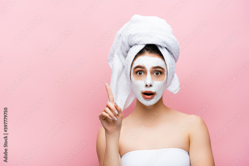 shocked beautiful woman with towel on head and clay mask on face pointing aside isolated on pink