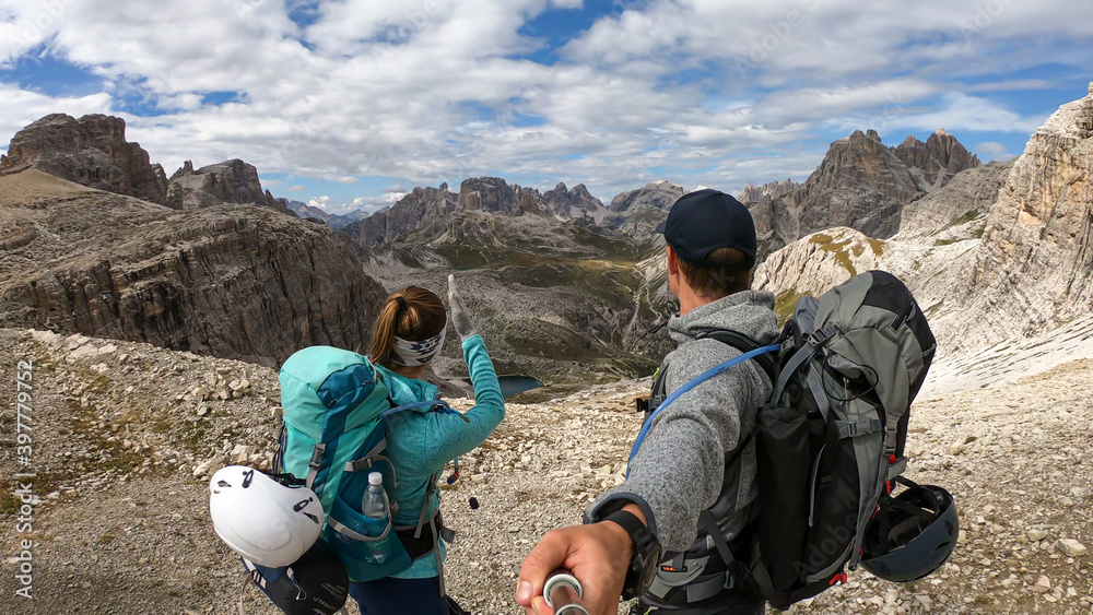 A couple with backpacks and sticks hiking on a narrow path in Italian Dolomites. There are sharp and steep mountains around. Lots of lose stones. Raw and desolated landscape. Following the pathway
