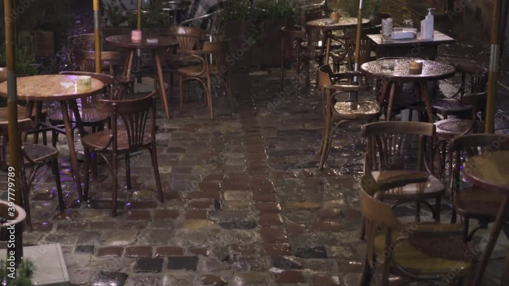 Old street cafe at night in the rain. Wet wooden furniture tables and chairs full of puddles. Raindrops fall on wet cobblestone street shimmering lights. Old European outdoor cafe in the rain Old street cafe at night in the rain. Wet wooden furniture tables and chairs full of puddles. Raindrops fall on wet cobblestone street shimmering lights. Old European outdoor cafe in the rain