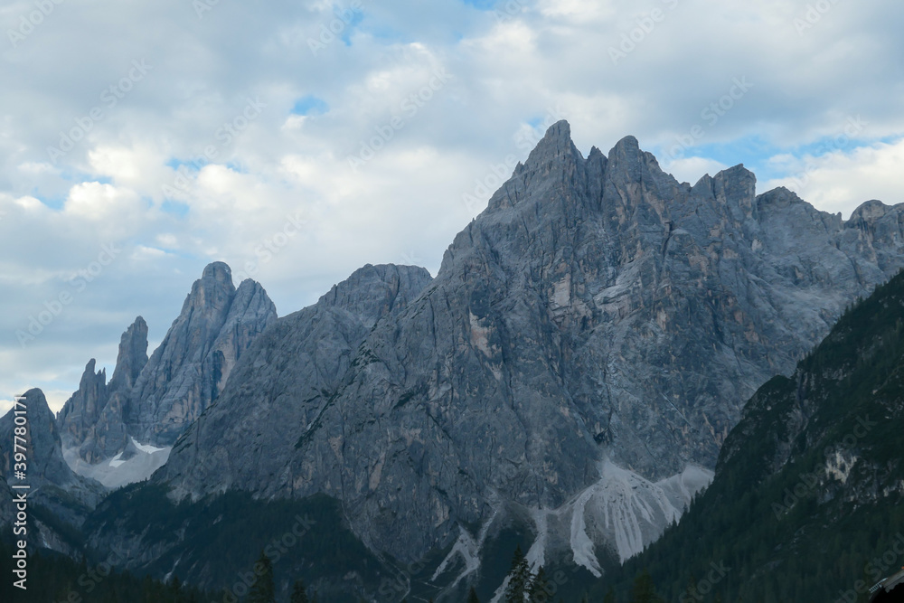 A close up view on the high and sharp peaks in Italian Dolomites. The slopes have a lot of landslides and lose rocks. Dense forest below the Dolomites. Few clouds above. Natural landscape