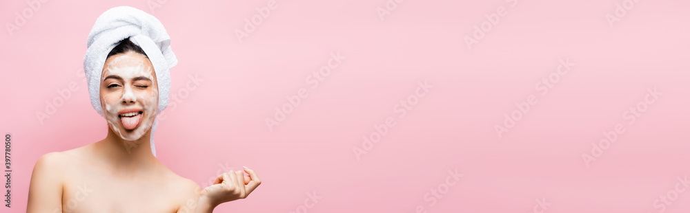 beautiful woman with towel on hair and foam on face showing tongue isolated on pink, banner
