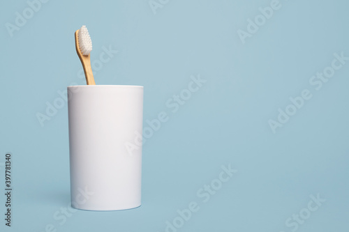 One wooden natural bamboo toothbrush in a white cup on a blue background with copy space and room for text with a left side composition