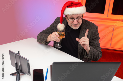 Remote Christmas party. Man in a Santa hat and a glass of champagne communicates in online conference. Man wearing Christmas hat drinking champagne and talking to friends on virtual zoom video call.