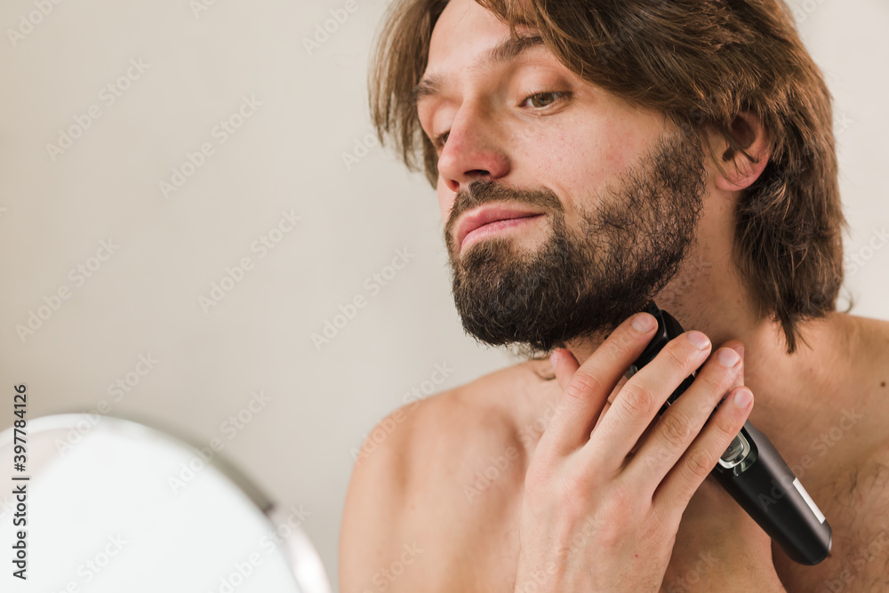 Obraz premium Man shaving his beard in the mirror with electric razor. Man with beard. Shaving his beard in the mirror with electric razor, indoors, coronavirus isolation concept and lockdown, new normal.