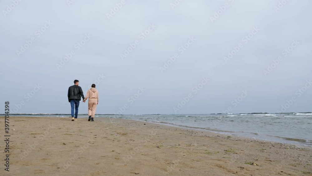 Young couple walks joining hands along empty Caspian sea beach past rolling waves under cloudy sky on nasty autumn day backside view Young couple walks joining hands along empty Caspian sea beach past rolling waves under cloudy sky on nasty autumn day backside view