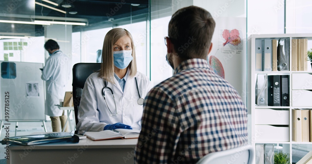 Rear of male patient sitting in cabinet in hospital on consultation with senior female doctor. Woman physician in medical mask talking and showing medicine to patient. Treatment concept. Covid-19 Rear of male patient sitting in cabinet in hospital on consultation with senior female doctor. Woman physician in medical mask talking and showing medicine to patient. Treatment concept. Covid-19