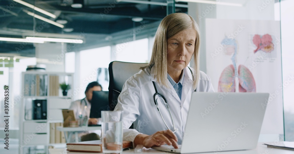 Portrait of beautiful senior busy Caucasian woman doctor sitting at desk in hospital office working and typing on laptop at workplace. Healthcare worker during coronavirus. Job concept