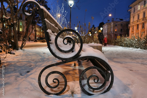 Winter night landscape , bench covered with snow among frosty winter trees and street lights. bench with black ornate cast iron legs. outdoor furniture