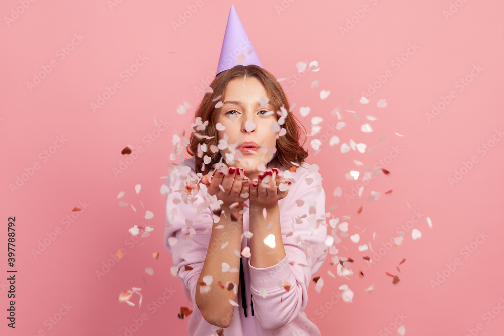 Portrait of carefree cheerful teenage girl in hoodie and party cone blowing heart shaped confetti, enjoying birthday or valentines day, festive mood. Indoor studio shot, isolated on pink background