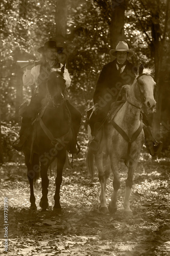 Vintage photo of two cowboy men riding on horses holding guns in a forest.