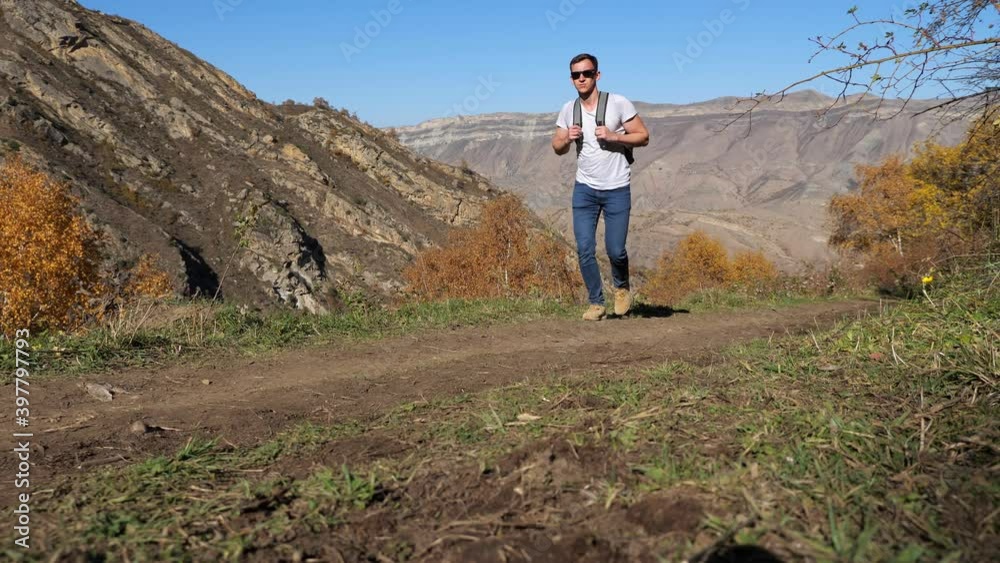 Young man tourist with sunglasses and backpack walks along narrow rural road near large ancient mountain against blue sky on nice autumn day Young man tourist with sunglasses and backpack walks along narrow rural road near large ancient mountain against blue sky on nice autumn day