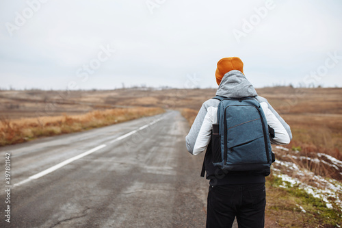 Male tourist walks on the side of an empty winter road in suburban area with a backpack. A man traveller wearing orange hat on a highway. All seasons tourism. Hitch-hiking, travelling concept. © Konstantin Zibert