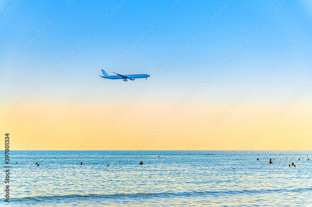 Airplane flying low above sea and people tourists swimming in water, clear blue orange sky at sunset, plane preparing to land at Larnaca airport above Mediterranean sea