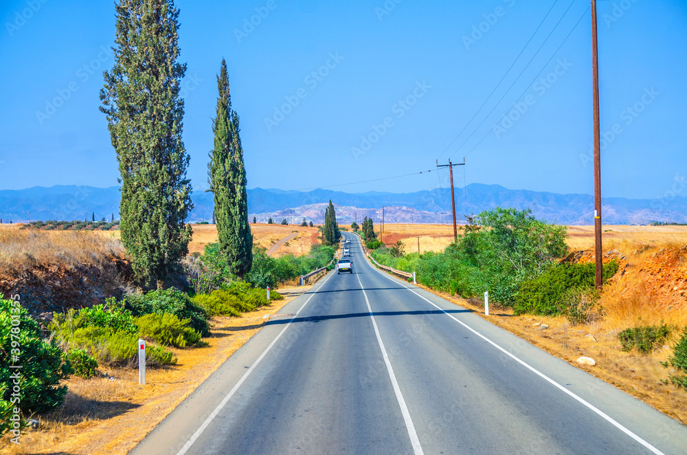 Fototapeta premium Landscape of Cyprus with cars vehicles riding asphalt road in valley with yellow dry fields, cypress trees and roadside poles, Troodos mountain range and hills, clear blue sky in sunny day background
