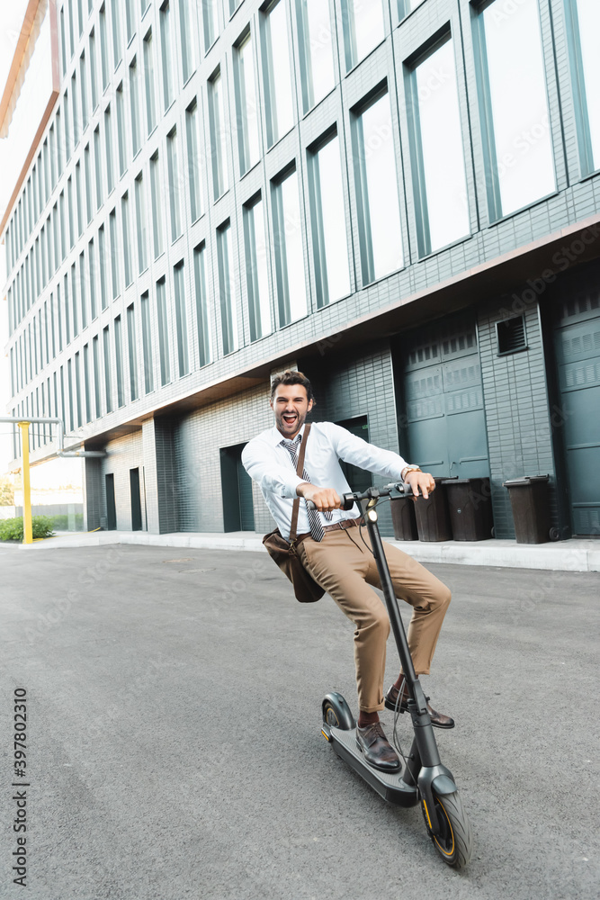 full length of excited businessman in formal wear riding electric scooter near building